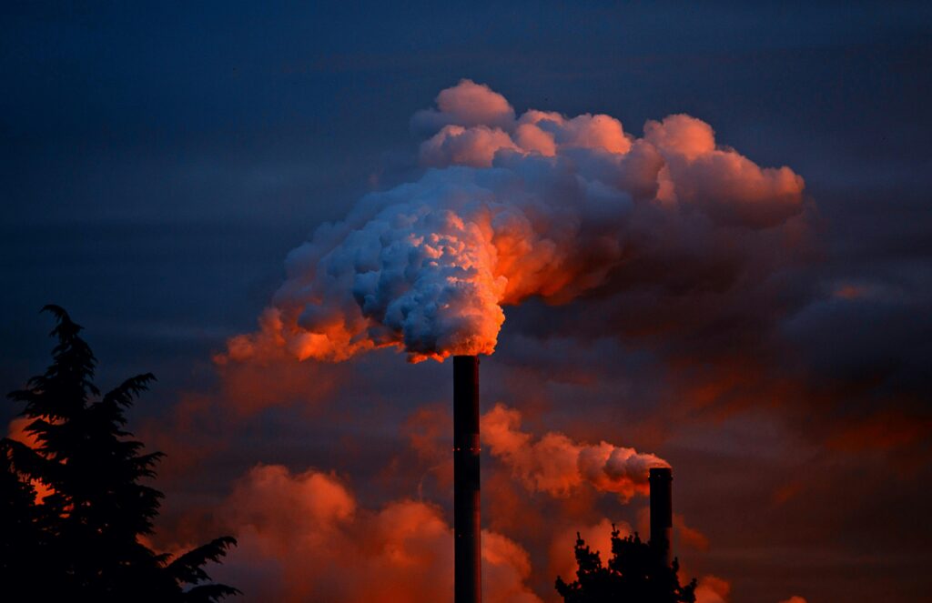 Smoke billows from industrial chimneys at sunset, highlighting pollution against a vibrant sky.